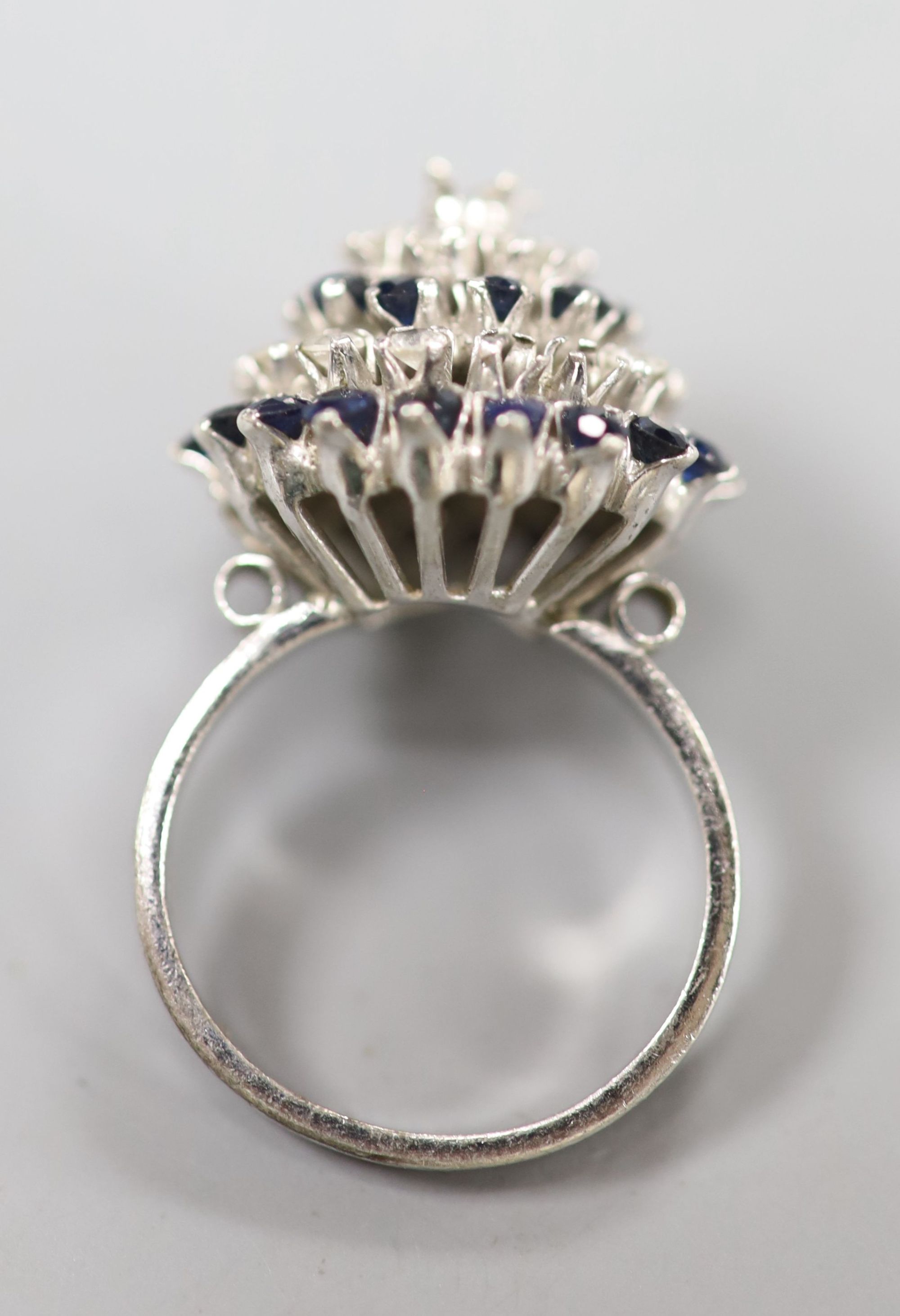 A white metal (stamped 18k) and two colour sapphire? set raised cluster ring, size K/L, gross 5.2 grams (one stone missing).